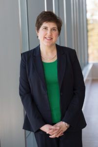 A woman in a suit and green shirt is standing in front of a window.