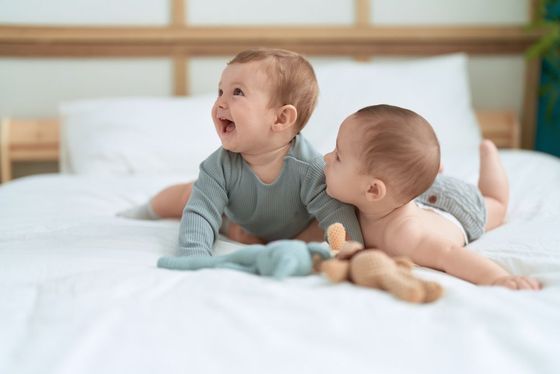 Two babies are laying on a bed looking at each other.