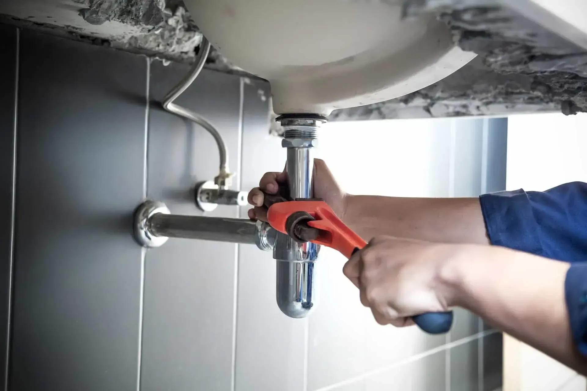 Plumber using a wrench to repair a sink's drain pipe in a bathroom with dark tiled walls.