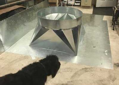 Metal ventilation component sits on a metal sheet. A black dog is in the foreground.