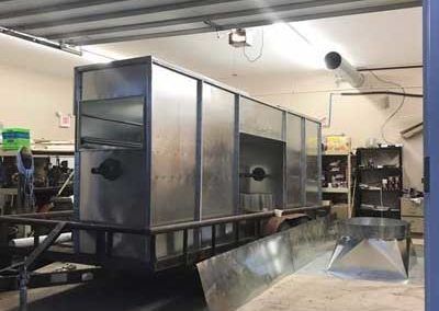 A large, metal rectangular structure on a trailer, possibly a smoker or oven, in a workshop.