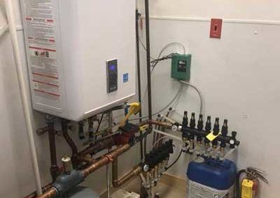 A white water heater with copper pipes, connected to a green control box and other plumbing in a utility room.