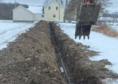 Trench dug in snow-covered field with an excavator, near a house.