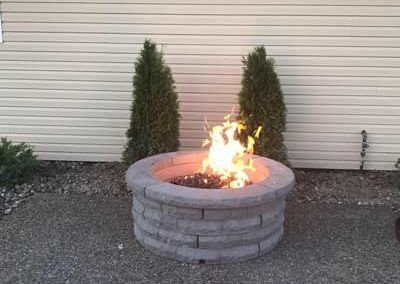 Fire pit with flames, flanked by evergreens, against a light-colored siding wall.