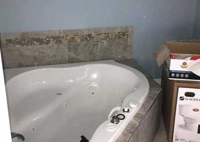 A corner whirlpool bathtub with partially tiled surround in a bathroom. A box sits next to it.