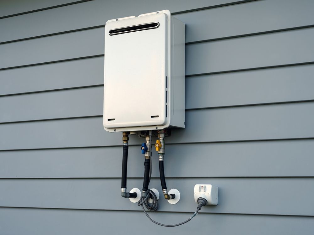 White tankless water heater mounted on gray siding. Black pipes and a white outlet are also visible.