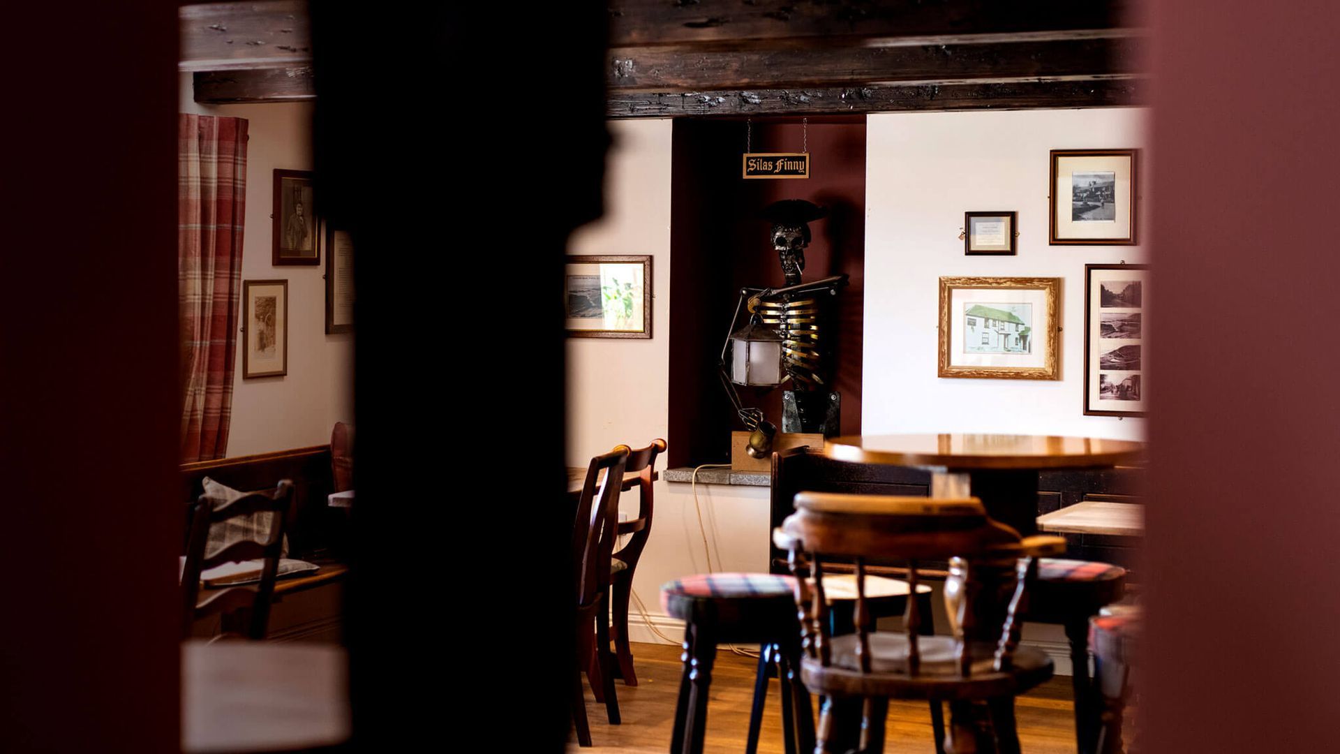 Interior view of a pub with wooden tables, chairs, framed art, and a dark wooden beam in the foreground.