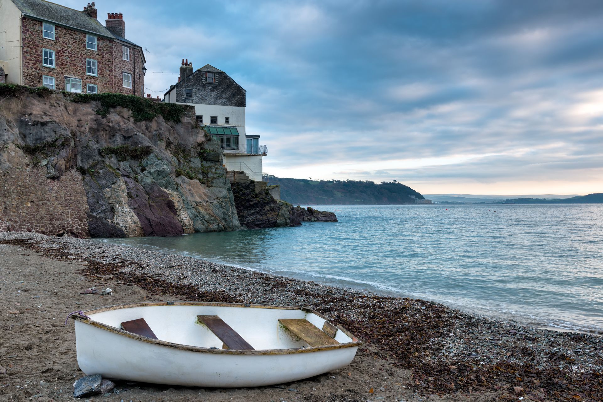 A white rowboat on a pebble beach with buildings atop a cliff overlooking the sea. Overcast sky.