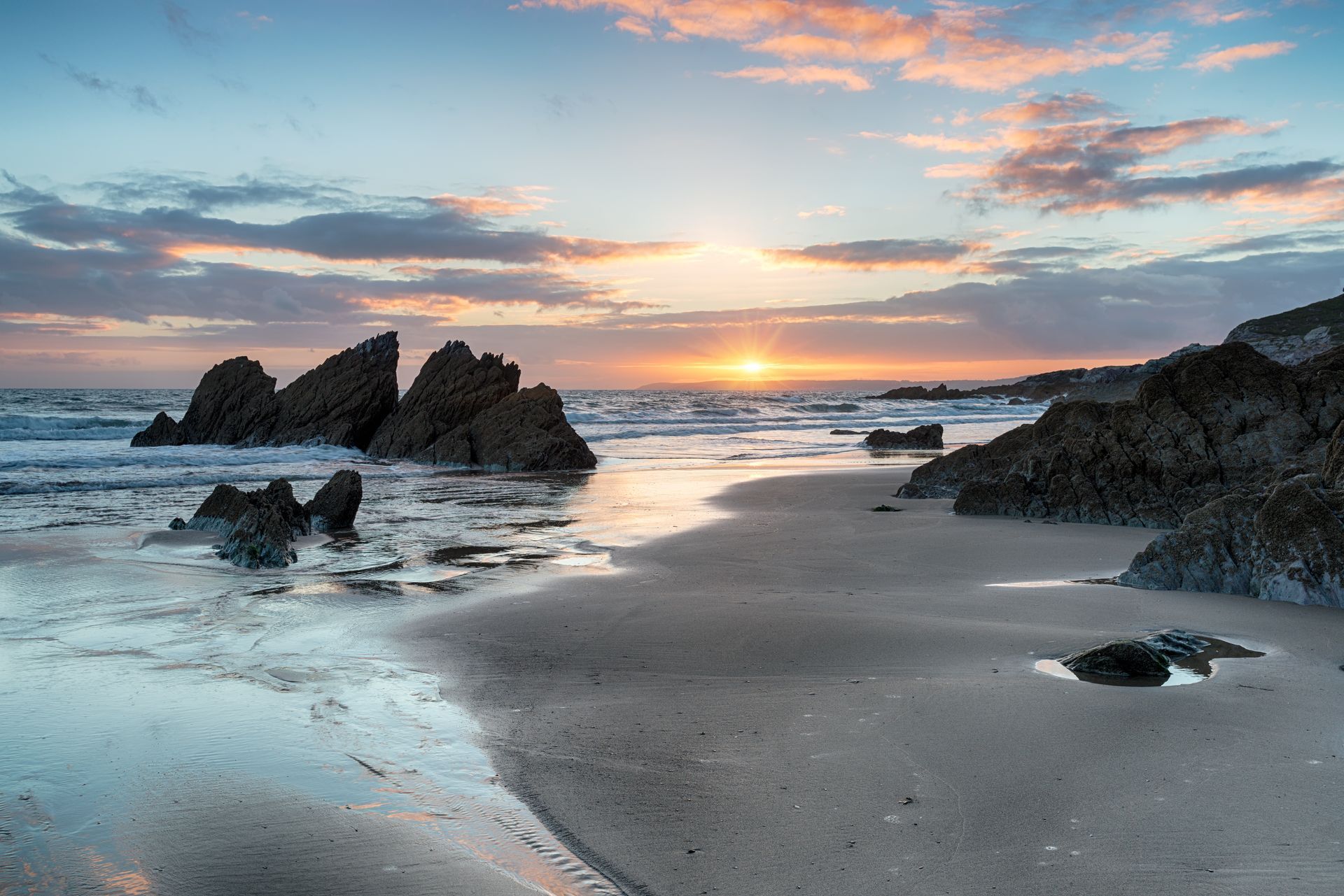 Beach at sunset, with wet sand, rocky outcroppings, and colorful clouds over the ocean.