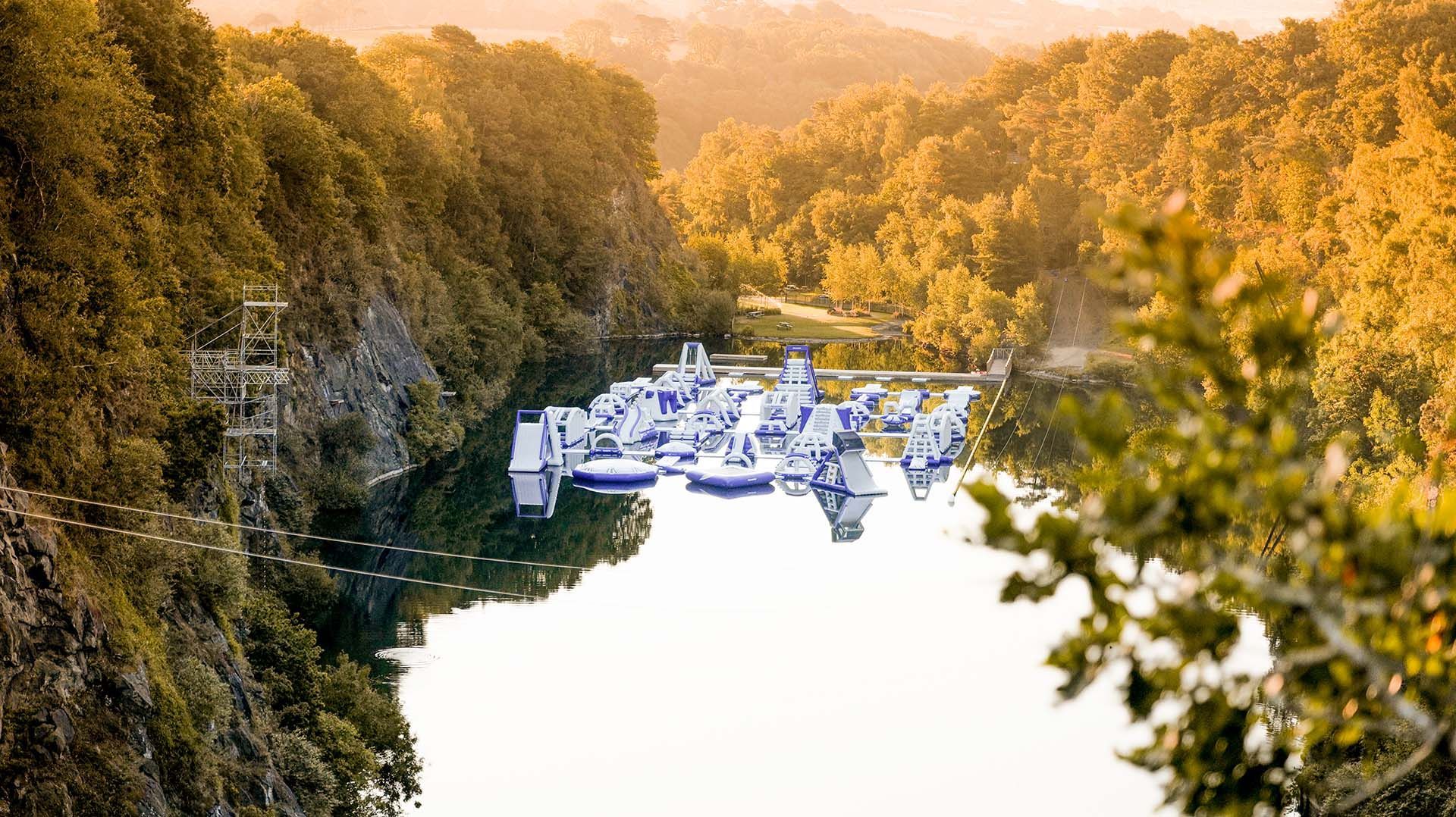 Water park with inflatable structures on a lake surrounded by cliffs and trees. Warm sunlight.