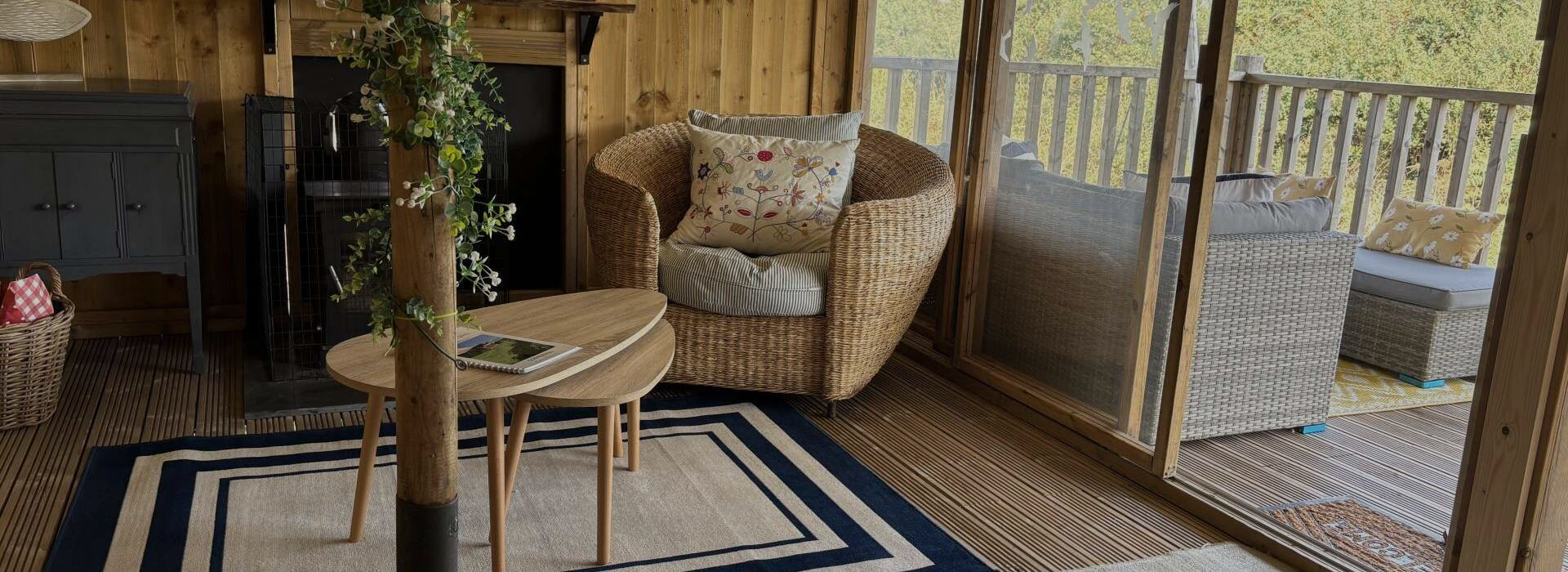 Cozy cabin interior with a wicker chair, coffee table, rug, and large windows looking out to nature.