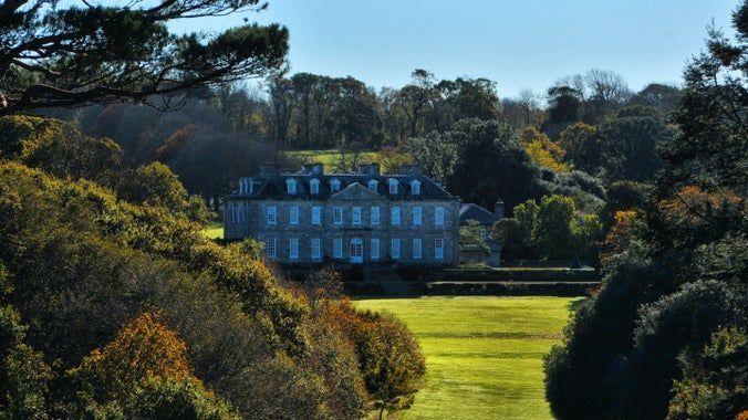 Stone manor house viewed from a tree-lined meadow on a sunny day.