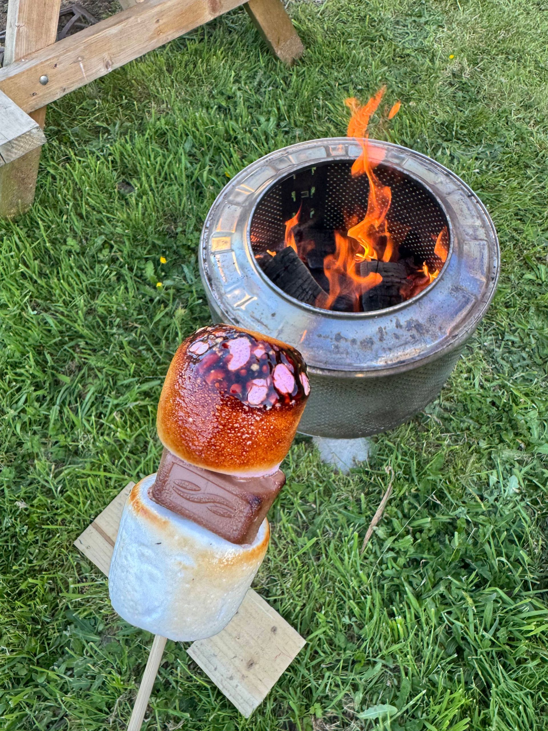 A s'more with toasted marshmallow and chocolate near a fire pit with flames.