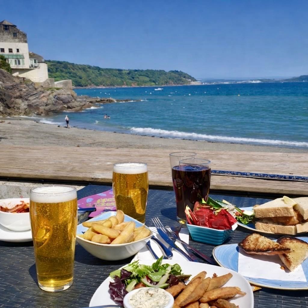 Outdoor table with food and drinks overlooking a beach and ocean. Beer, fries, salad, sandwiches, sunny day.
