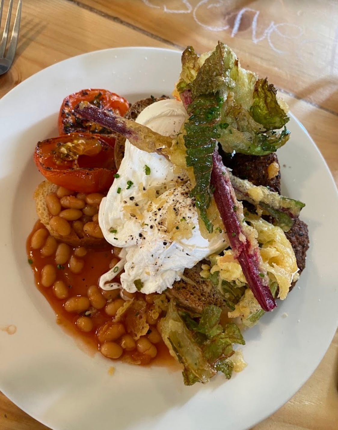 White plate with a breakfast spread: poached egg, baked beans, tomato, fried greens, and what looks like fried bread.