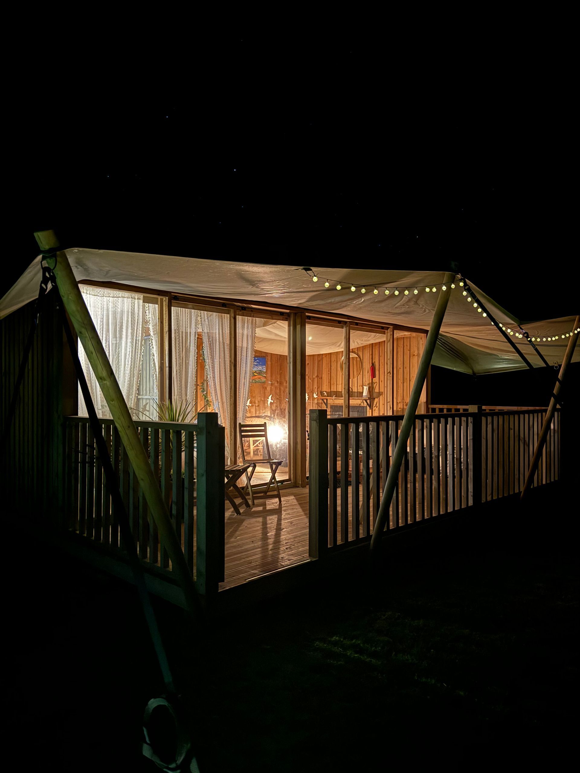 Wooden deck with canopy lit by string lights; view into cabin with furniture, at night.