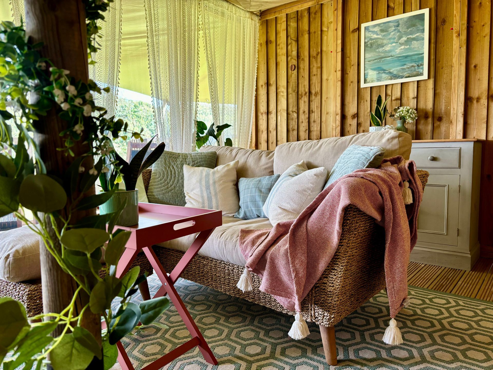 Cozy cabin interior: sofa with pillows and blanket, plants, wooden walls, rug, and small side table.