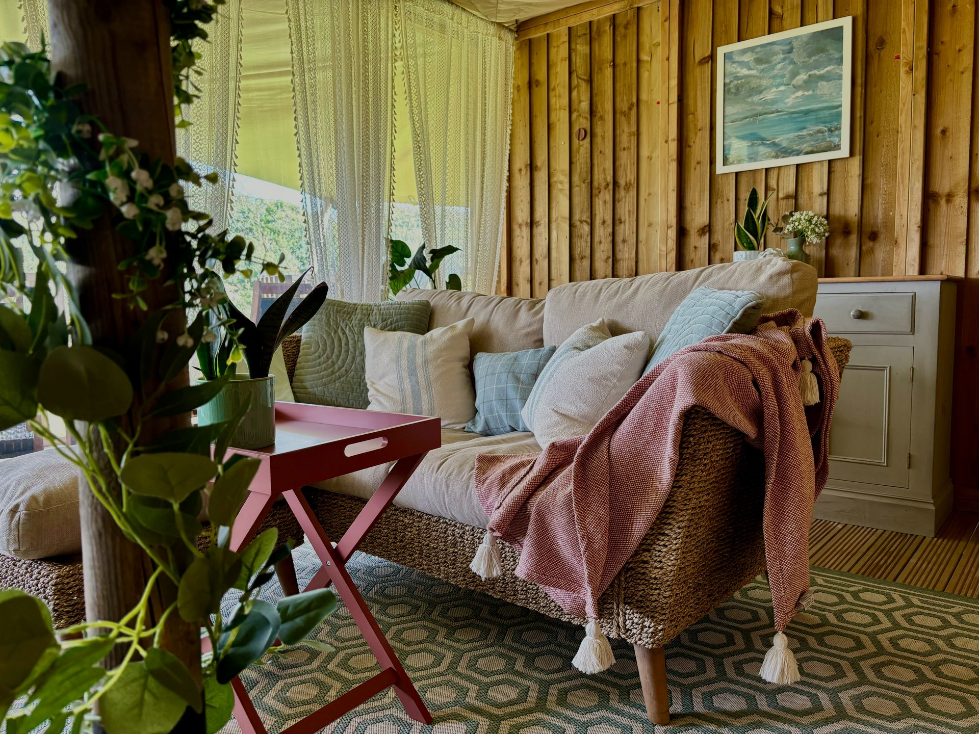 Cozy cabin interior: sofa with pillows and blanket, rug, wooden walls, framed art, and greenery.