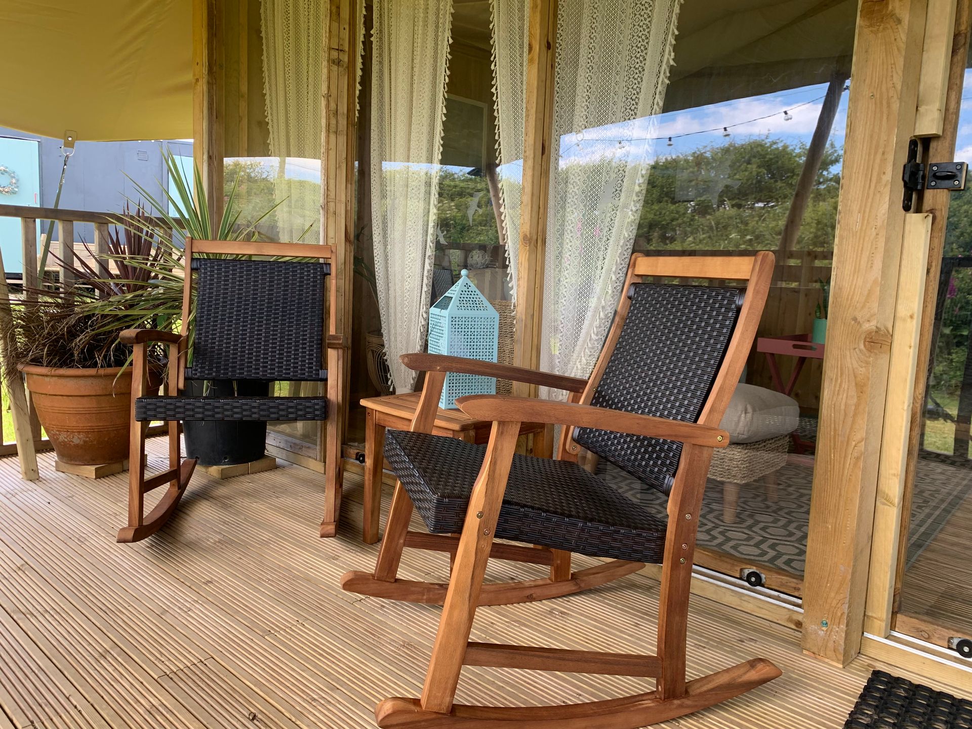 Two wooden rocking chairs on a porch with woven mats. Glass doors and sheer curtains reveal greenery.