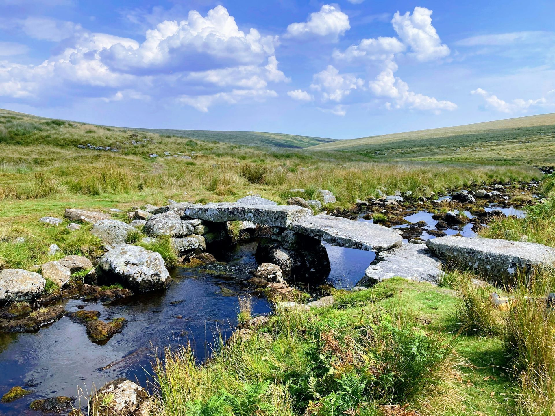 Stone bridge over a stream in a grassy valley under a blue sky with clouds.