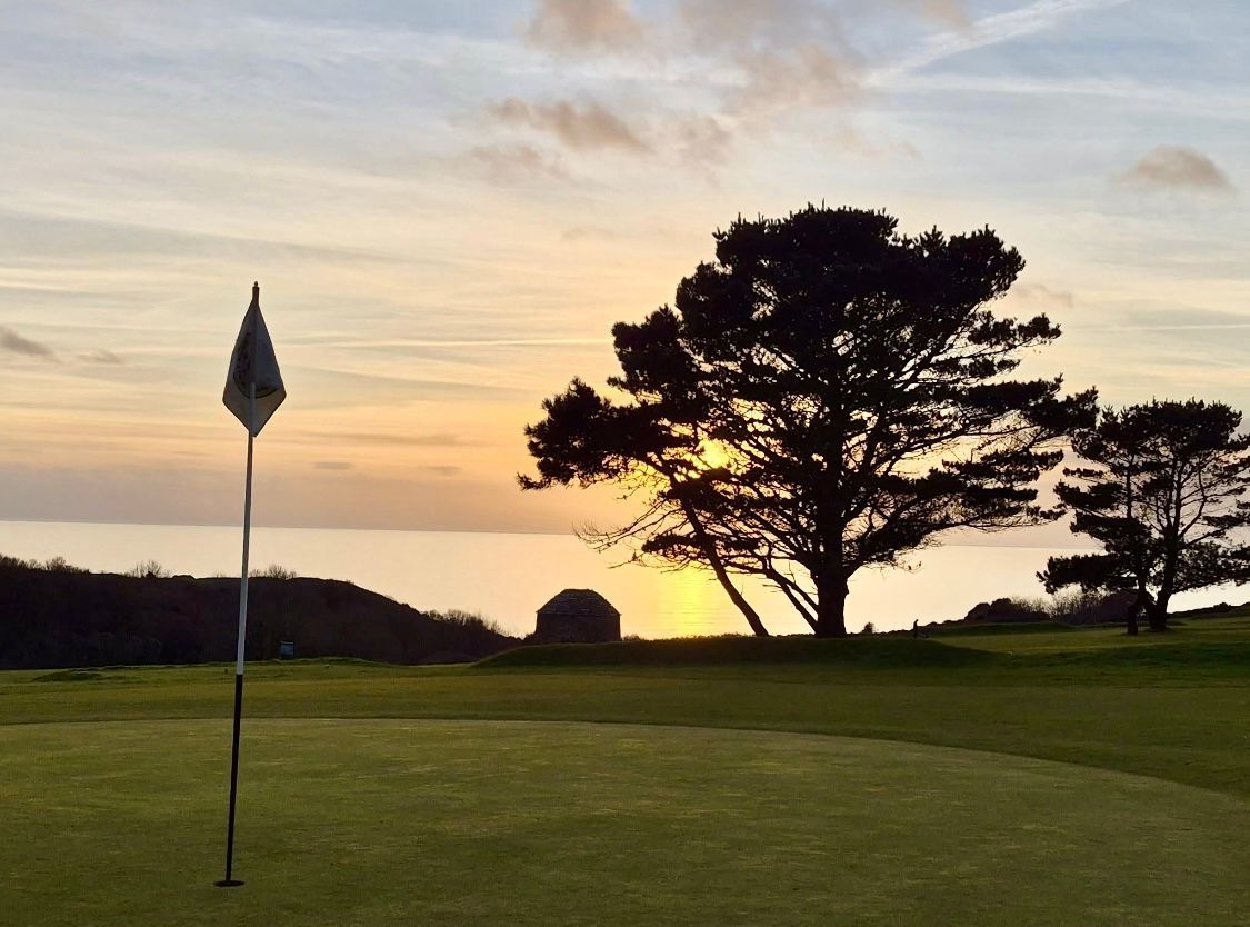 Sunset on a golf course: flag on green, trees silhouetted, ocean and sky in background.