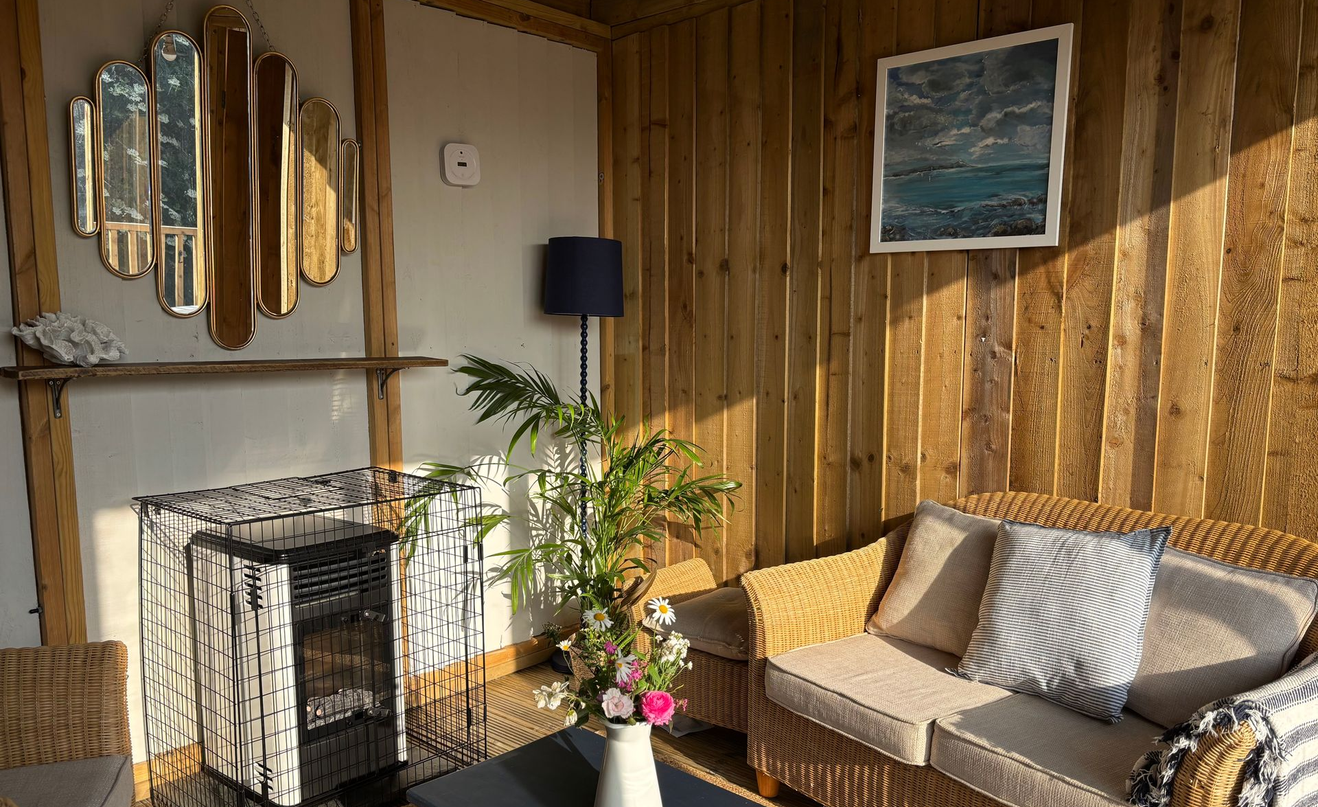 Cozy sunroom with wood paneling, wicker sofa, framed art, decorative mirror, and potted plants.