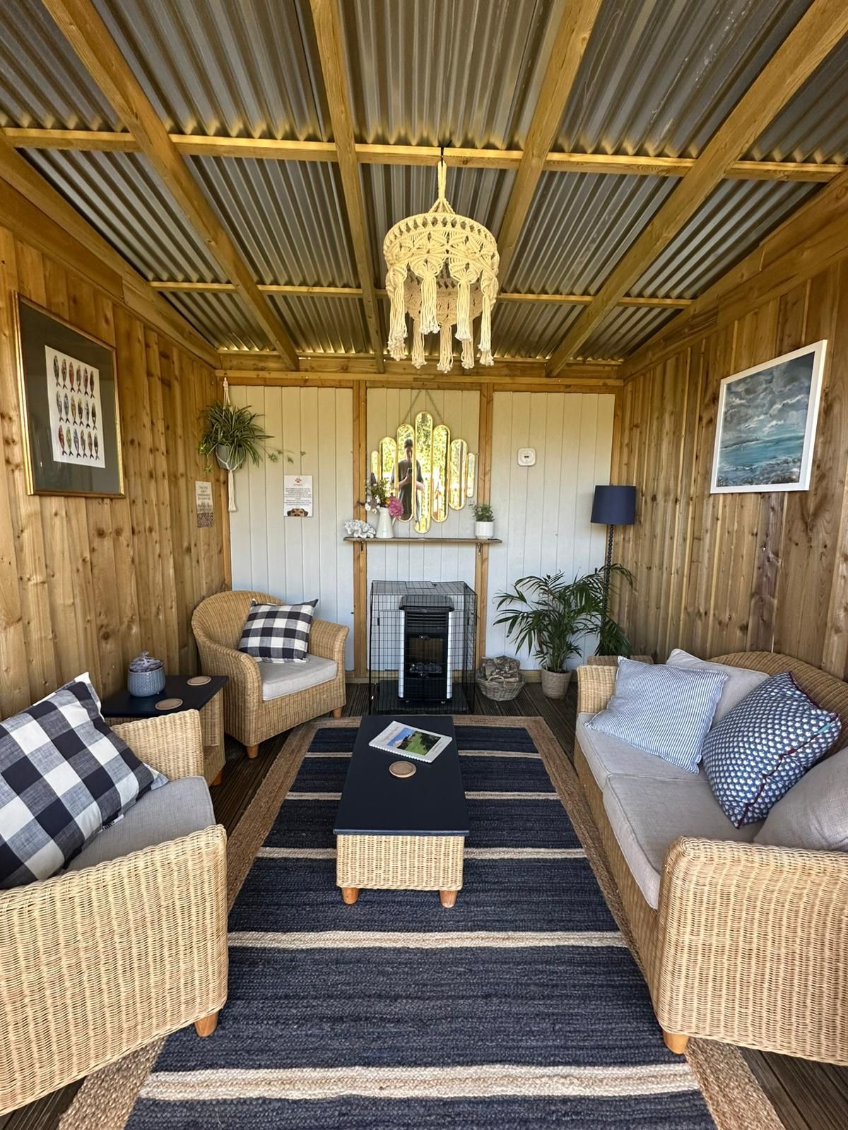 Cozy outdoor room with wicker furniture, a rug, and a macrame chandelier. Wooden walls, corrugated metal roof.