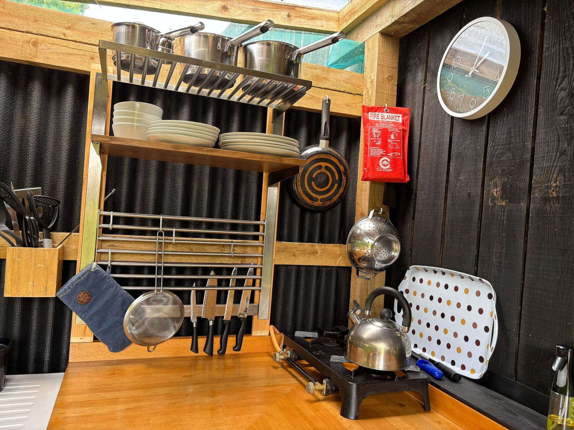 Small outdoor kitchen with pots, pans, utensils, and a gas stove on a wooden counter.