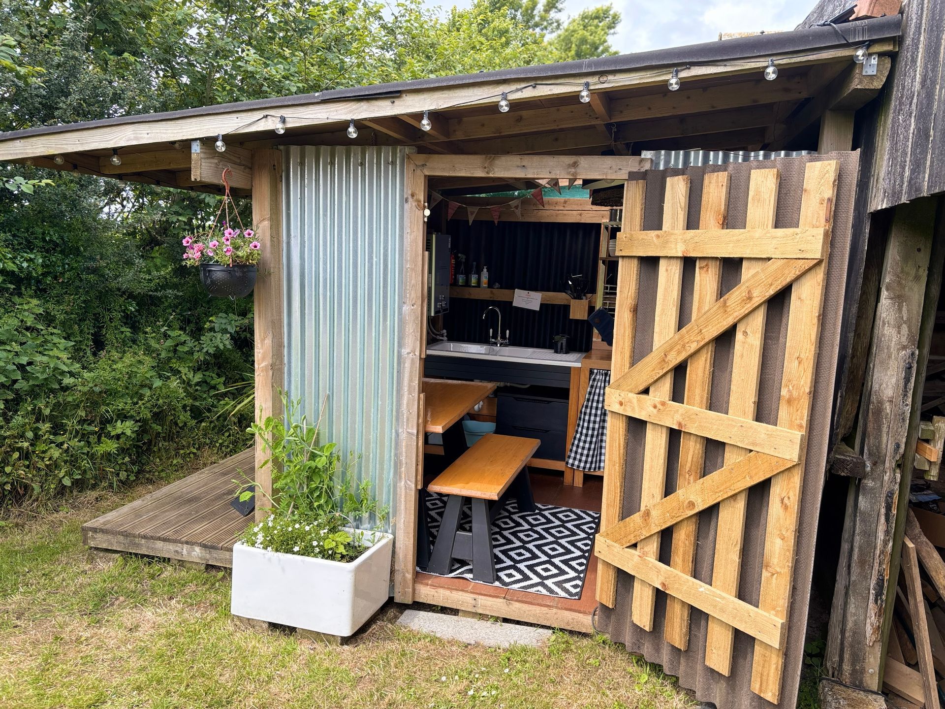 Rustic outdoor shed with corrugated metal wall and pallet door, open to reveal a small kitchen space.