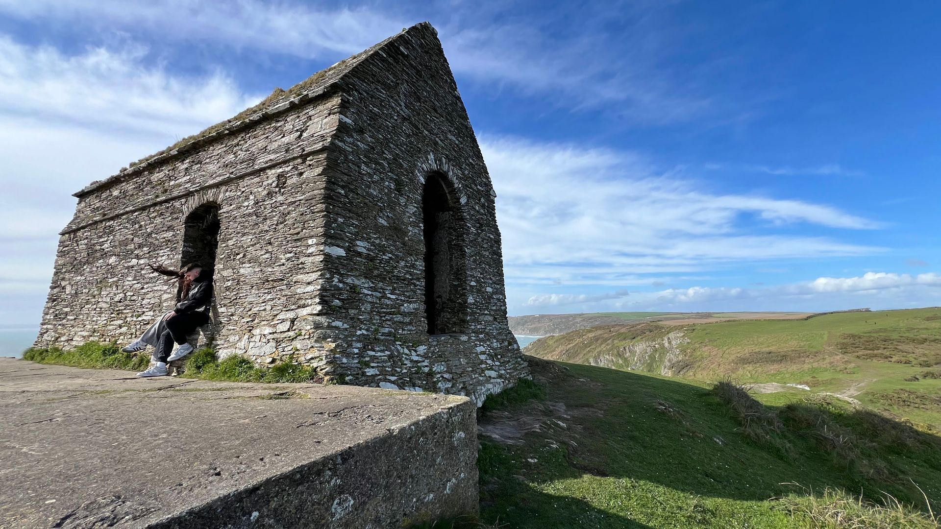 Stone structure on a grassy cliff overlooking a coastal landscape. A person sits in the doorway, blue sky above.