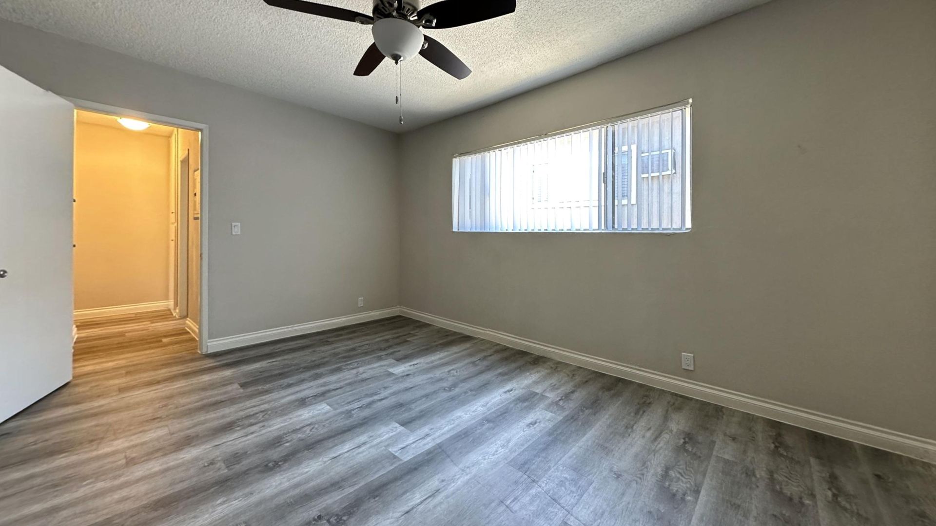 Empty room with gray walls, wood-look flooring, window with blinds, ceiling fan, and open door.