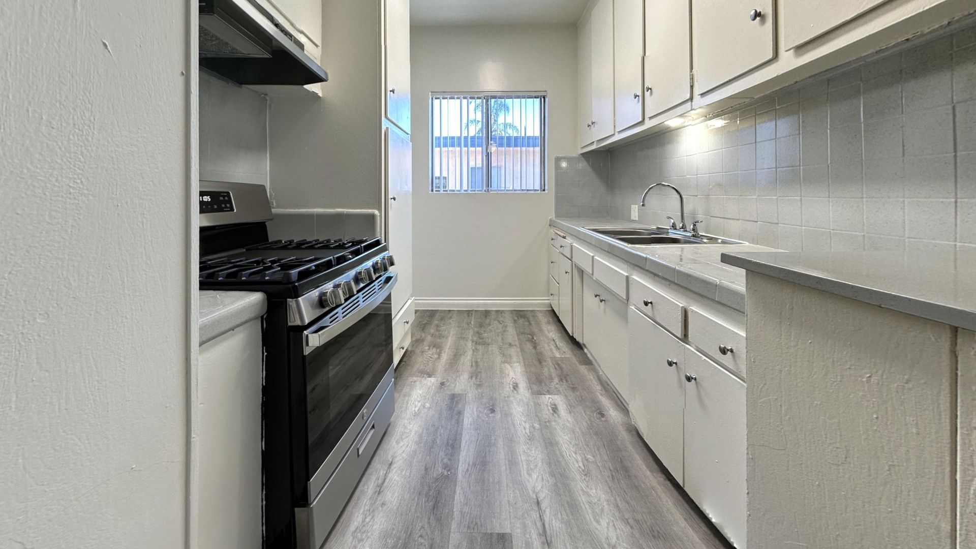 Narrow kitchen with stove, sink, and white cabinets. Gray flooring and a window at the end.