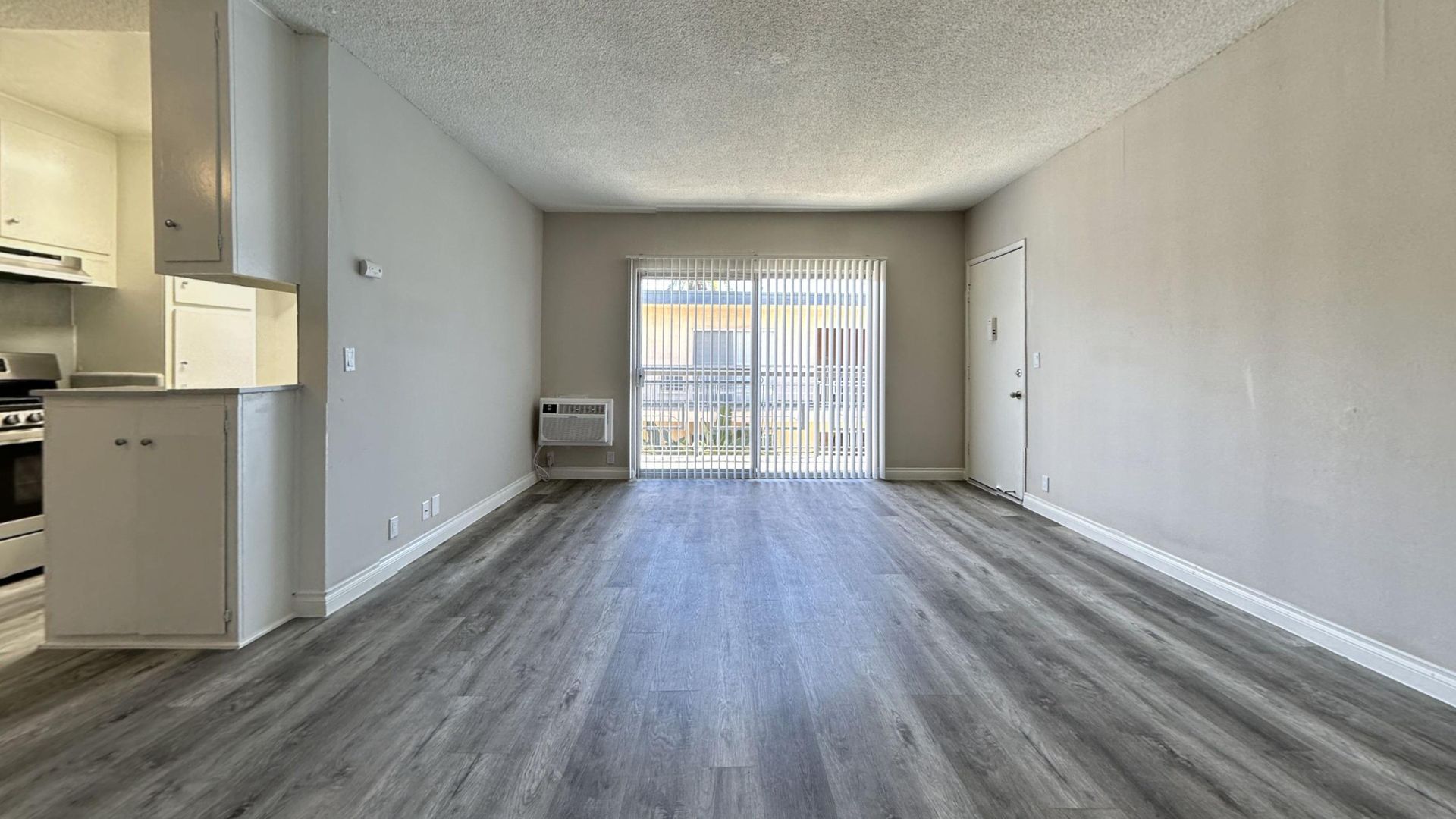 Empty apartment living space with gray laminate floors, sliding door, and kitchen.