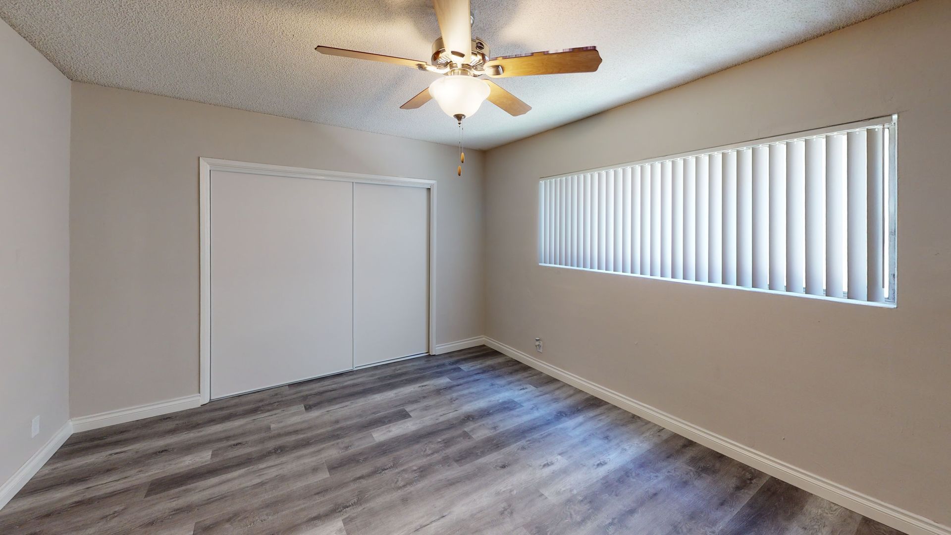 Empty room with gray laminate flooring, white walls, closet, and window with blinds. Ceiling fan present.
