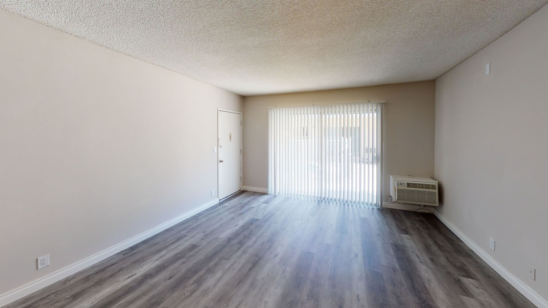 Empty apartment room with gray flooring, sliding glass door, and a wall-mounted air conditioner.
