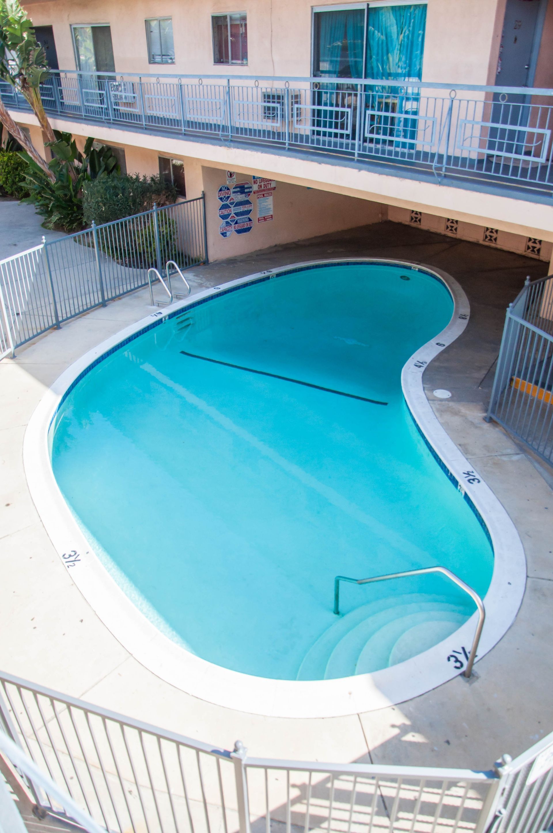 A turquoise, kidney-shaped swimming pool in a courtyard surrounded by a two-story apartment building.