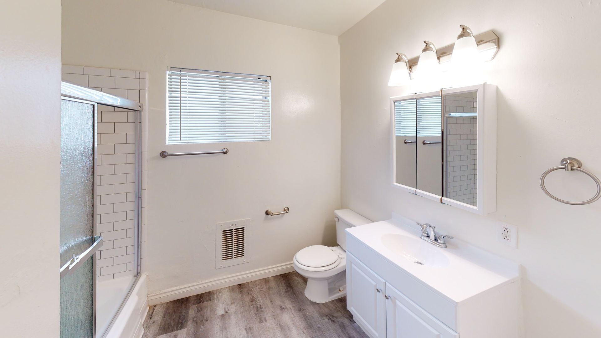 White bathroom with a shower, toilet, sink, and window. Gray wood-look floor.