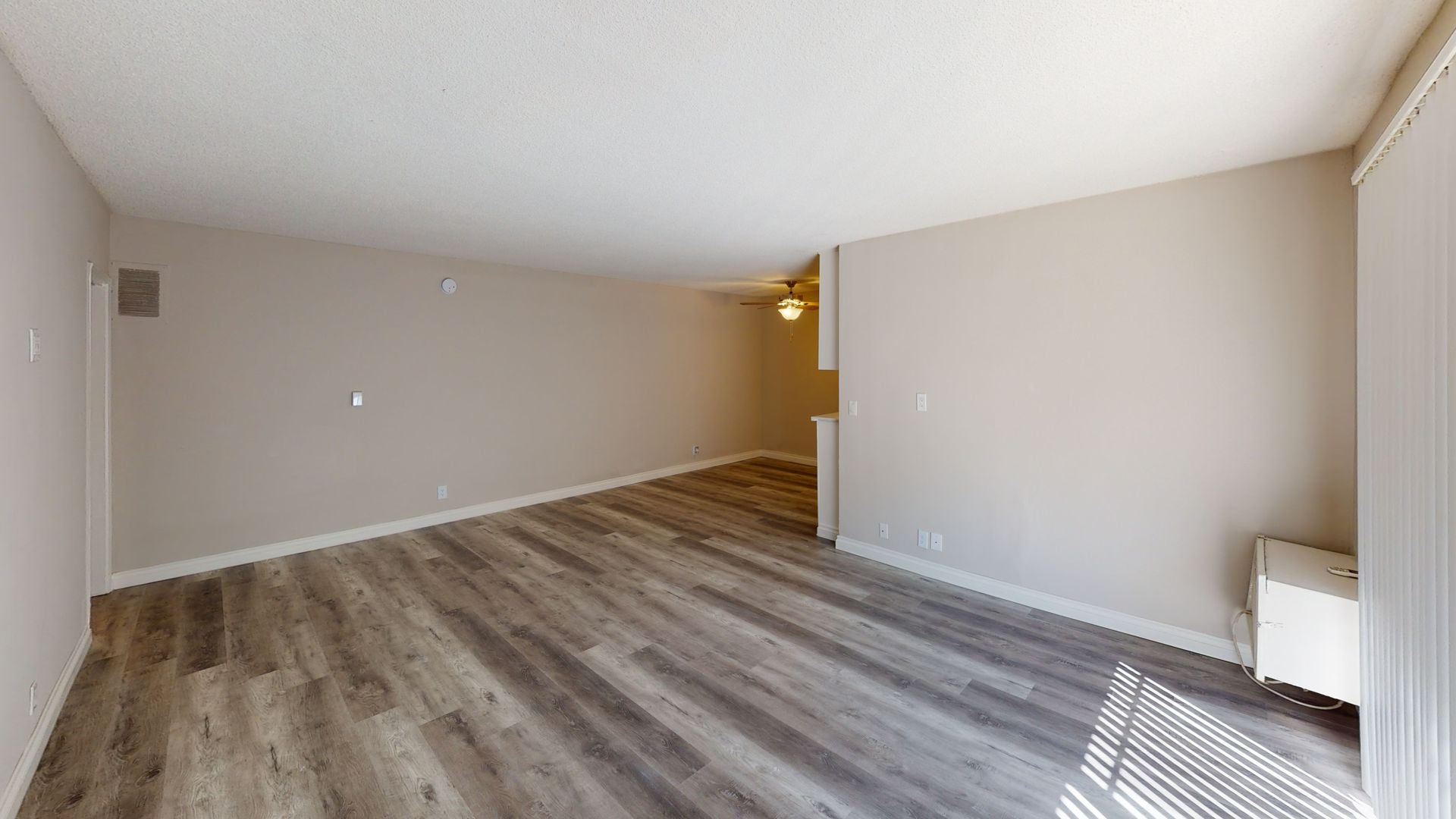 Empty living room with wood-look flooring and beige walls. White ceiling and blinds are on the right.