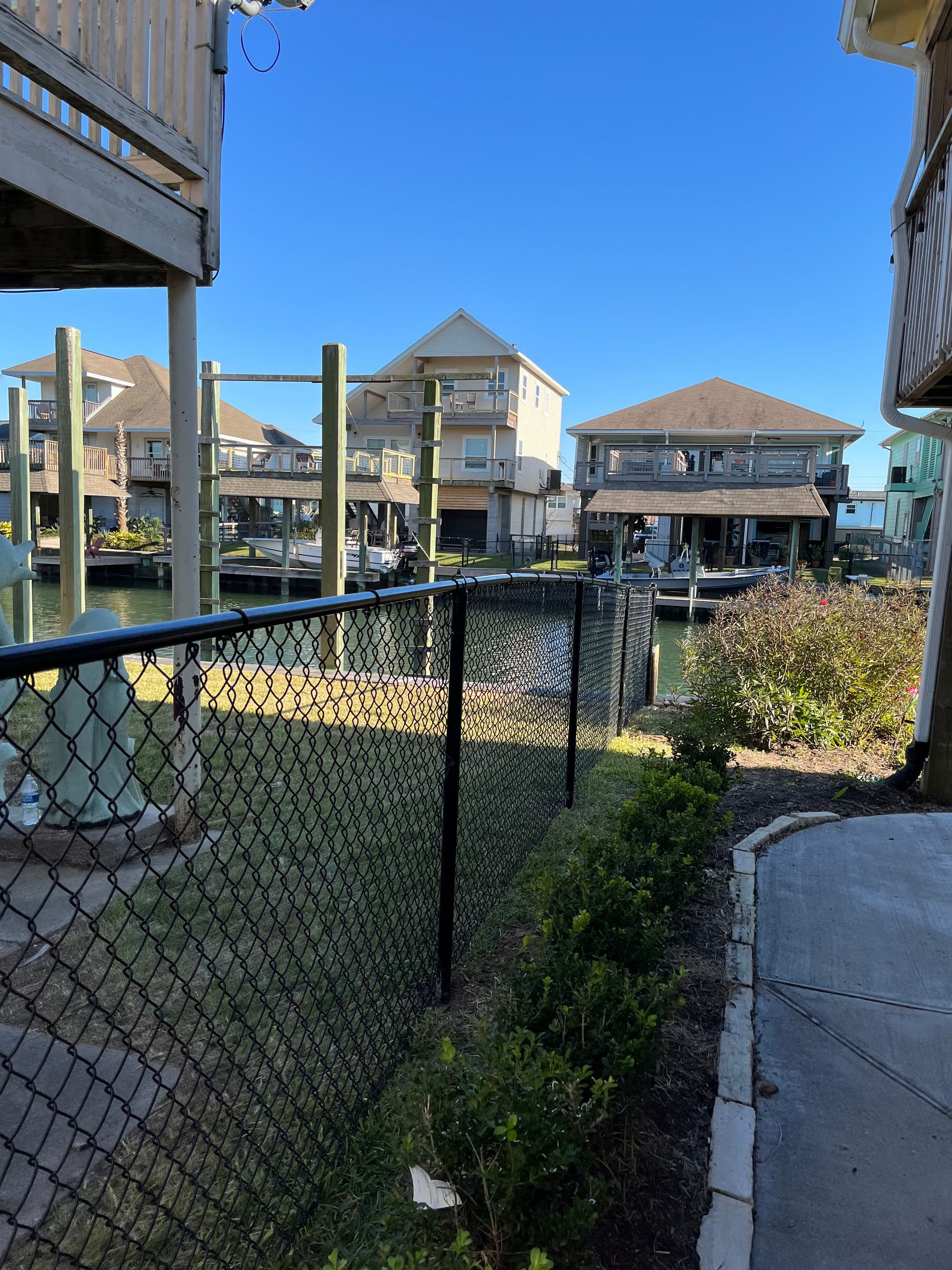 Waterfront homes with docks, seen from behind a black chain link fence on a sunny day.
