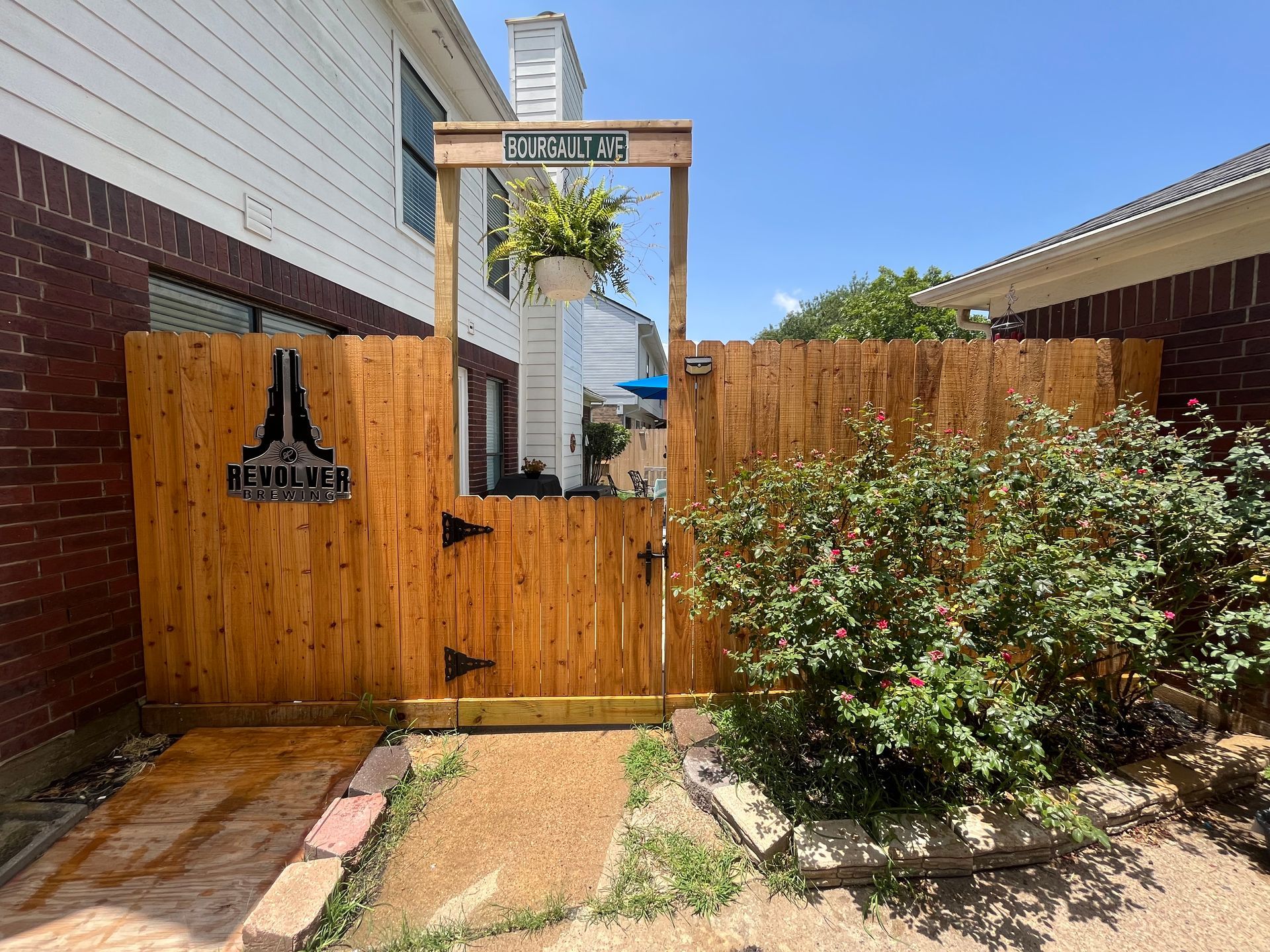 Wooden gate with a sign and a hanging plant, set between brick walls. Sunny outdoor scene.