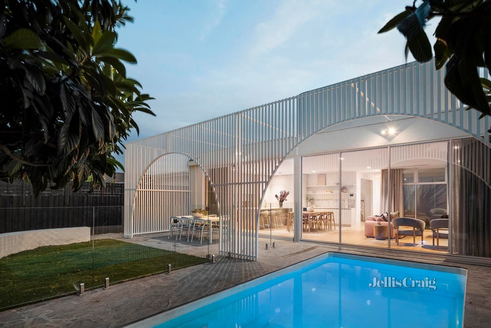 Modern house with slatted white facade beside a lit blue swimming pool at dusk