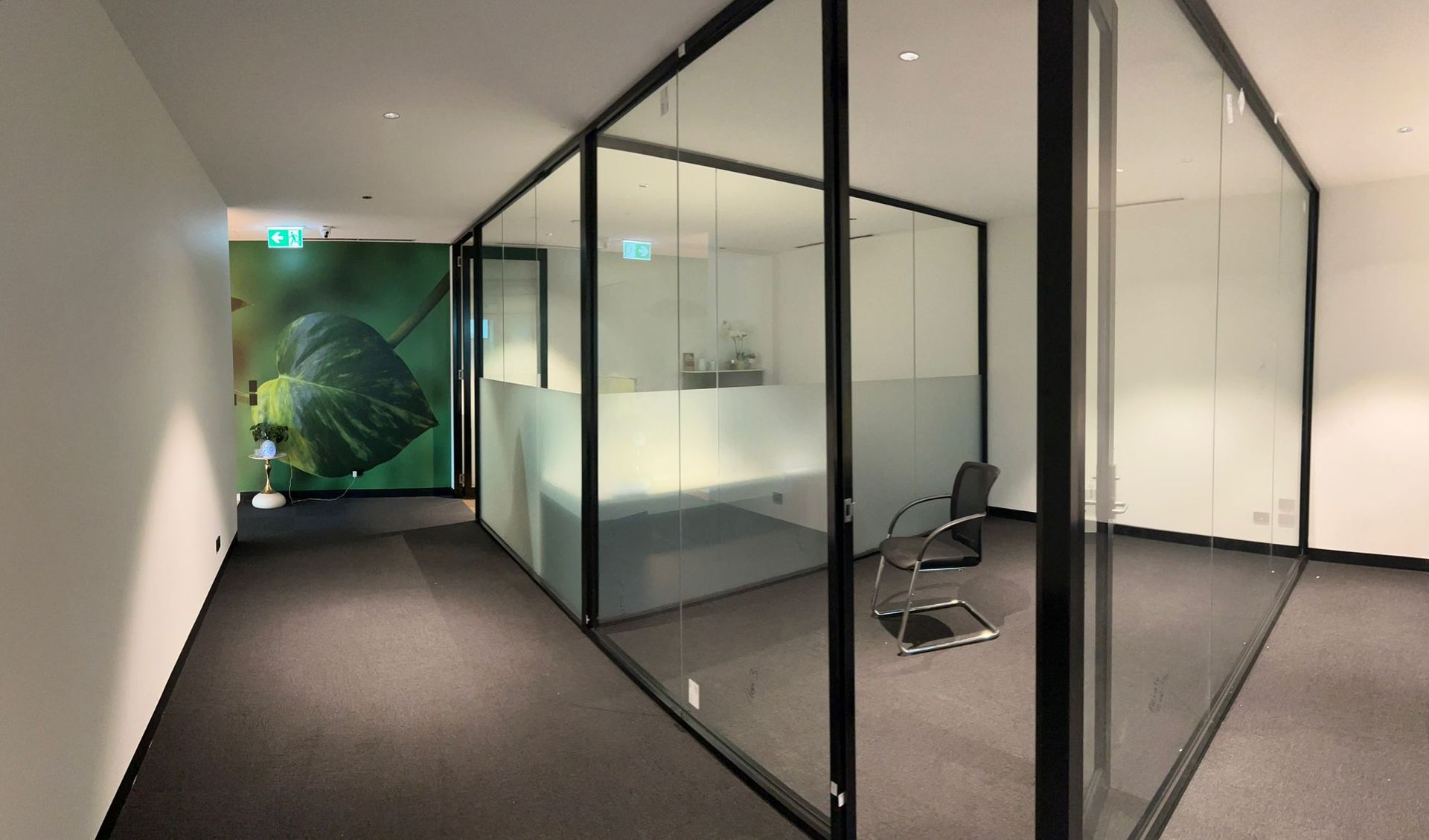 Modern office hallway with glass-walled meeting rooms and a few chairs on a dark carpeted floor