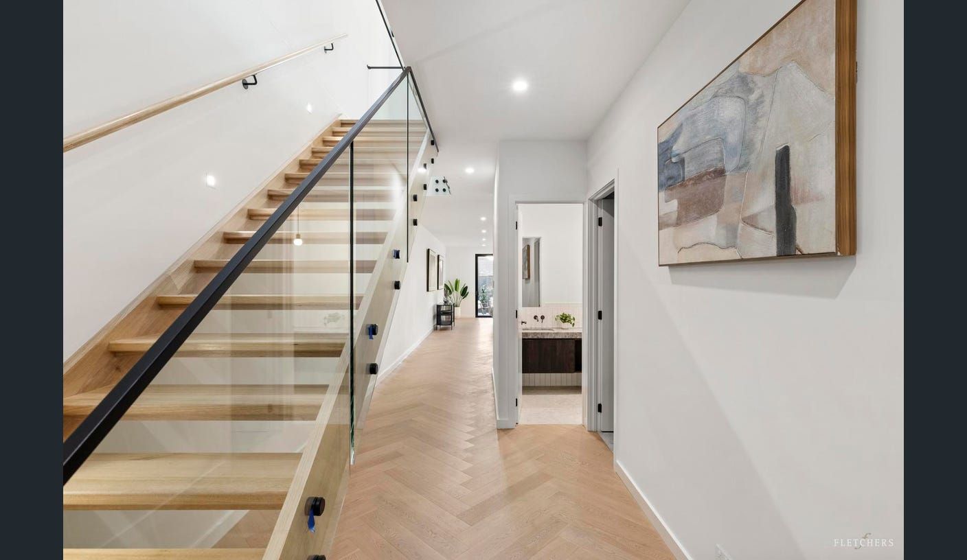 Modern hallway with glass staircase, herringbone wood floor, white walls, and framed wall art