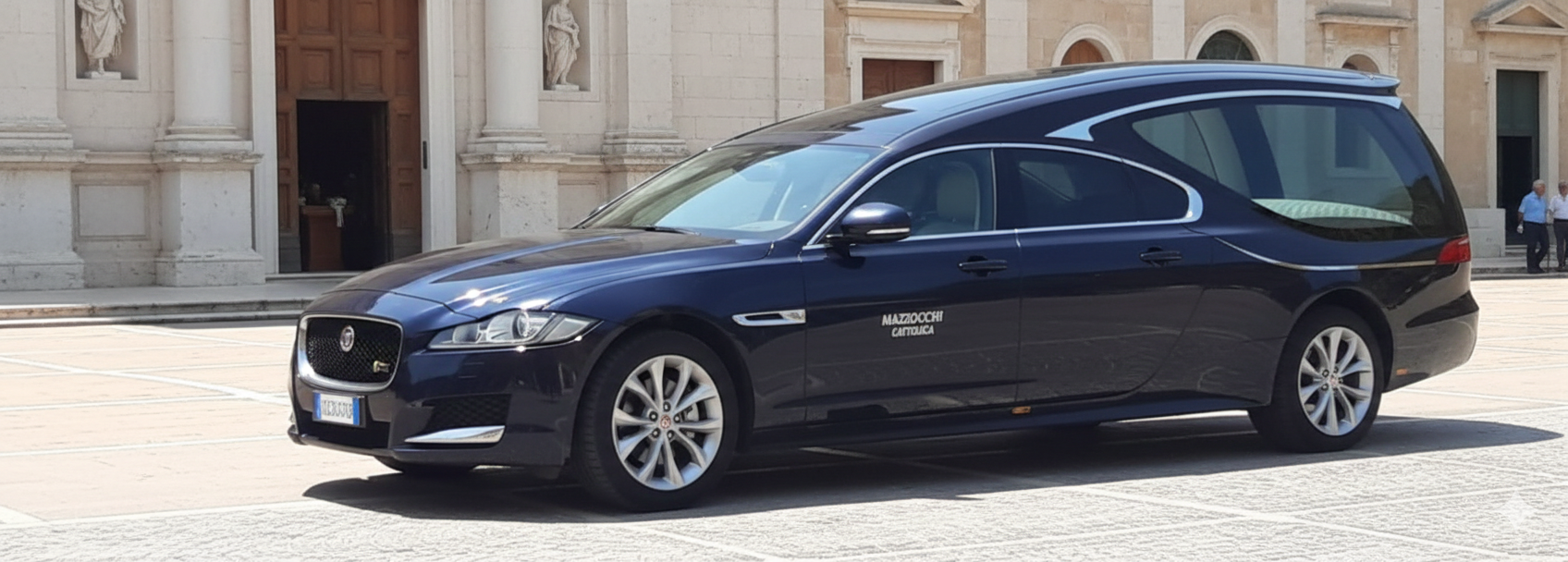 Un carro funebre Jaguar blu scuro parcheggiato davanti a un edificio con colonne e un grande portone.