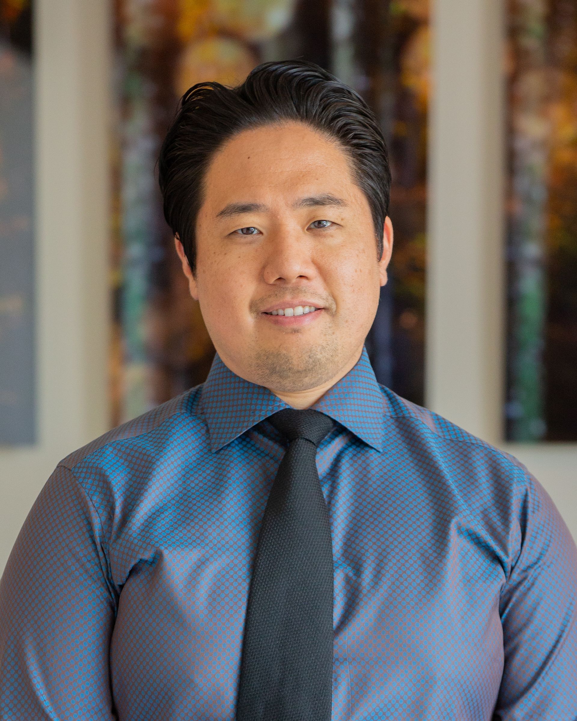 Man wearing a blue dress shirt and black tie smiling at the camera.