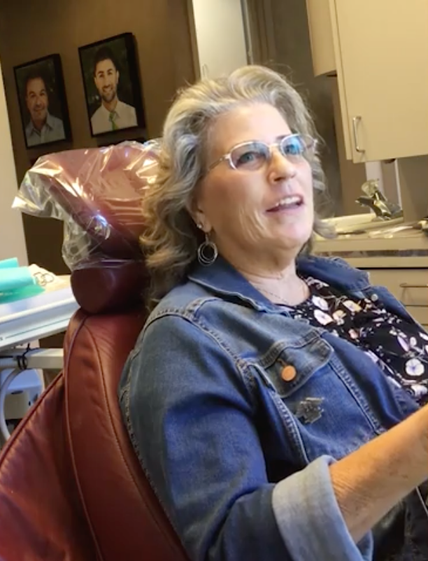 Woman wearing glasses and denim jacket in a dental chair; smiling, looking up. Dental office setting.