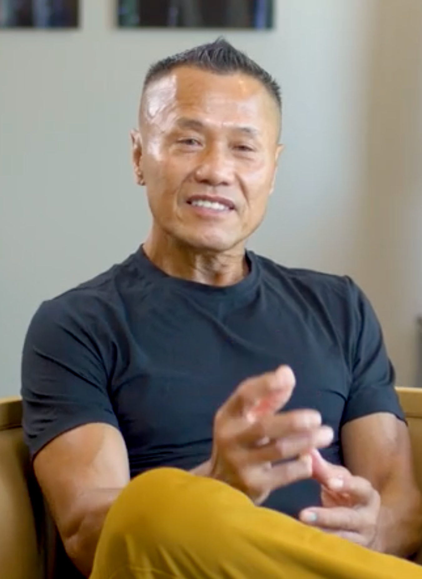 Man in black shirt, gesturing with hands, seated indoors, gold pants, short hair.