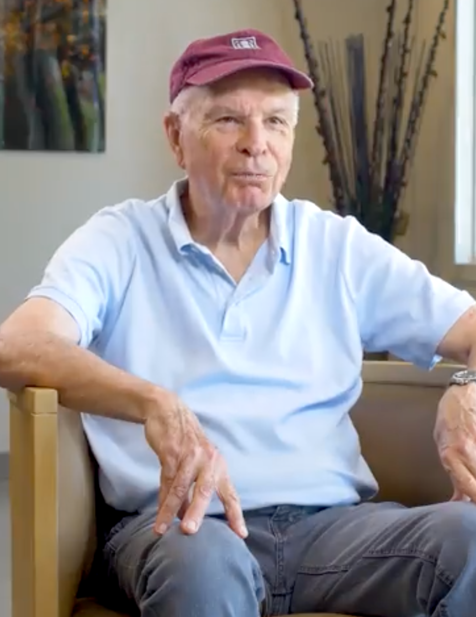 Older man in blue shirt and maroon cap sits indoors, looking forward.