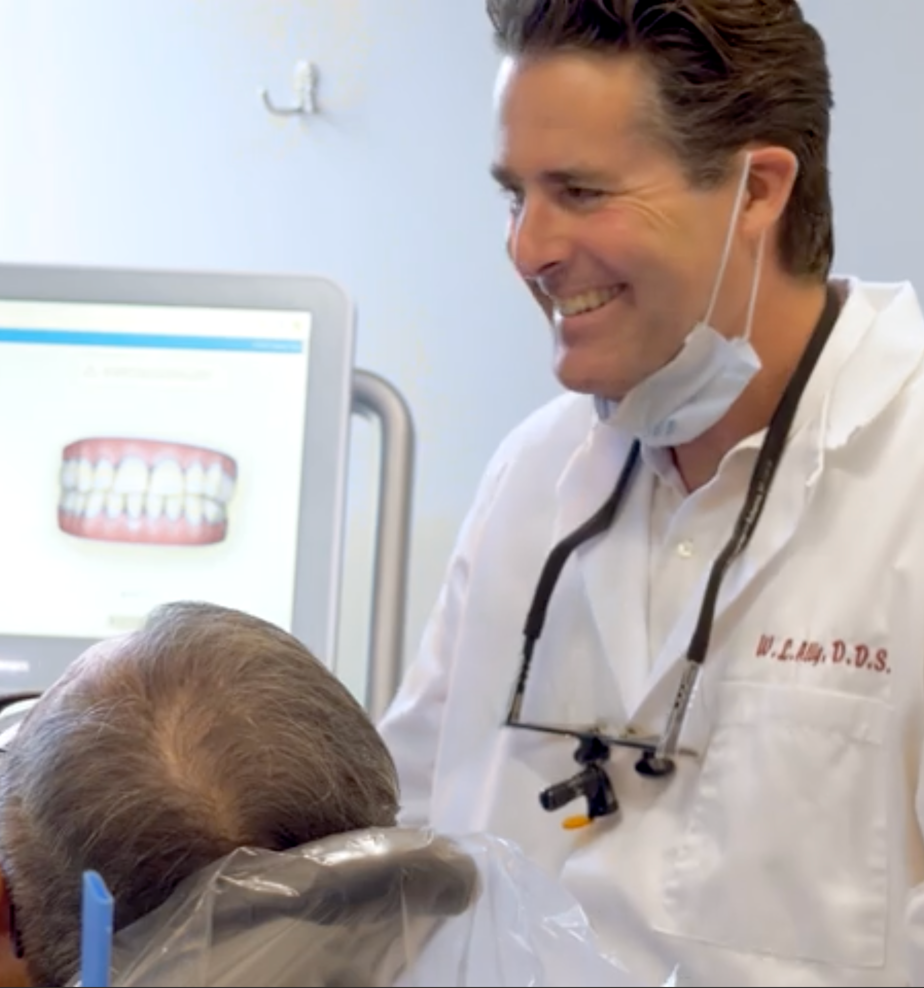 Dentist smiling, examining patient's teeth, computer with teeth image in background.