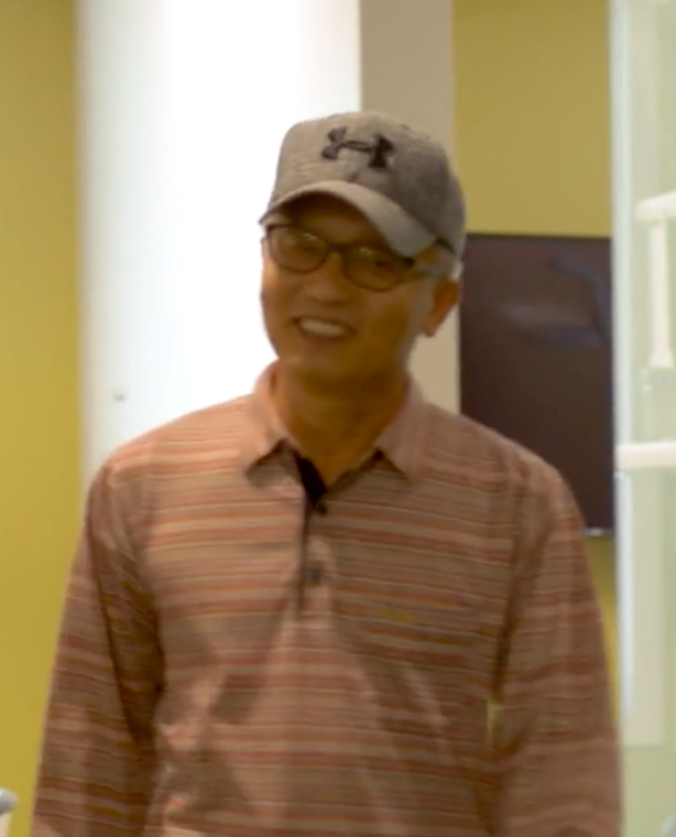 Man wearing a gray hat and glasses, smiling, wearing a striped polo shirt, standing indoors.